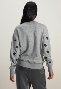 Sweat-shirt gris avec un col et des poignets côtelés, orné de motifs étoilés noirs sur les manches. Tissu doux avec une coupe décontractée.