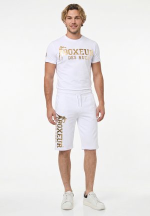 Junger Mann steht, trägt weißes T-Shirt und Shorts mit goldenem Schriftzug „Boxeur des Rues“ und weiße Sneaker vor weißem Hintergrund.