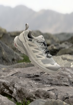 Chaussure de randonnée Salomon gris clair avec des attaches noires flottant au-dessus d'un terrain rocheux avec des montagnes floues en arrière-plan.