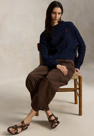 Μπλε ναυτικό πουλόβερ με πλέξη καλωδίου και ριμπ τελείωμα, καφέ παντελόνι σε χαλαρή γραμμή και καφέ σανδάλια με κυκλικά μεταλλικά διακοσμητικά, όλα πάνω σε ουδέτερο φόντο.