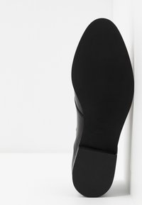 Semelle de chaussure habillée en cuir noir tournée vers l'avant, reposant contre un mur et un sol blanc dans un fond minimaliste propre.