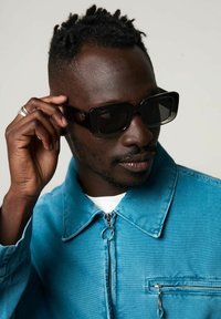 Lunettes de soleil noires carrées avec des montures épaisses, portées par une personne dans une veste en denim bleu vif avec une poche zippée, sur un fond neutre.