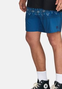 Shorts de bain bleus avec un imprimé géométrique en bleu plus foncé, dotés d'une taille contrastante. Le matériau semble léger et lisse.