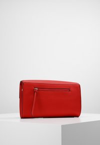 Borsa a tracolla in pelle rossa con una tasca con zip e un lungo tirante, appoggiata su un piedistallo bianco su uno sfondo grigio semplice.