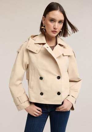 Kvinde iført beige croppet dobbeltradet jakke med sorte knapper, blå jeans og store guld- og brune øreringe mod en neutral baggrund.