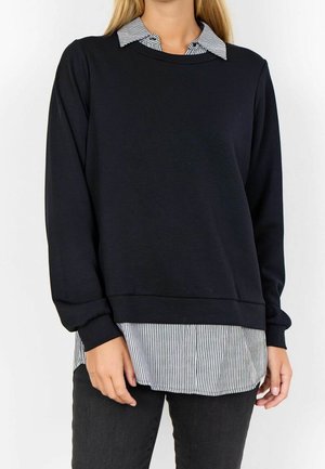 Vrouw draagt een zwarte trui met lange mouwen over een zwart-wit gestreept shirt met kraag, gecombineerd met donkere broek, staand tegen een witte achtergrond.