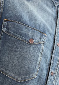 Denimskjorta med ljusblå färg, två bröstfickor, metallknappar och sömnadsdetaljer som ger en texturerad look.