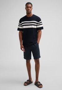 Maglione a maniche corte a righe navy con strisce orizzontali bianche, abbinato a pantaloni corti scuri e sandali neri con suole in sughero.
