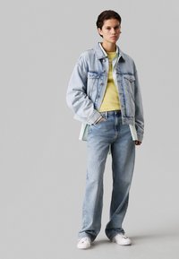 Spijkerjack in een lichte was met twee voorzakken, gecombineerd met een groen-geel shirt en lichtblauwe spijkerbroek. Model draagt witte sneakers.