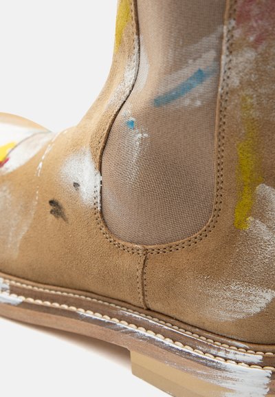 Μπεζ μποτάκι Chelsea από σουέντ με ελαστική πλευρική επένδυση, καφέ σόλα και σκόρπιες πιτσιλιές μπογιάς σε λευκό, κίτρινο, μπλε και μαύρο.