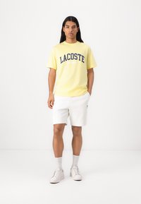 Gul bomulls t-skjorte med marineblå "LACOSTE" logo, kombinert med hvite shorts. Modellen har på seg hvite joggesko og sokker, stående mot en ensfarget bakgrunn.