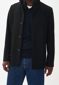 Svart kappa med rak skärning, knäppning framtill, hög krage och en slät yta. Bärs över en marinblå tröja och blå jeans.