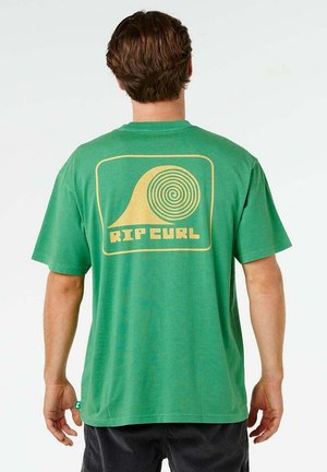 Homme portant un t-shirt vert Rip Curl avec un logo de vague en spirale jaune dans le dos, debout devant un fond blanc uni.