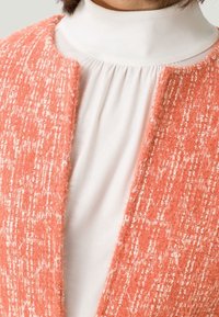 Koralfarvet cardigan med tekstur, fluffy overflade, åben front og ingen lukninger, båret over en hvid højhalset bluse med plisseringer.