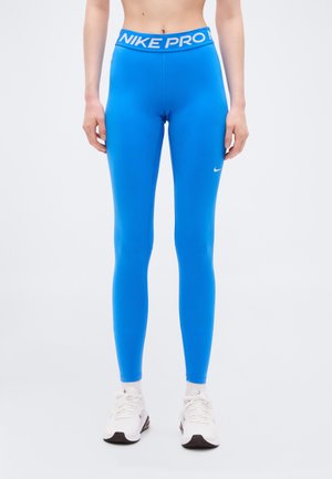 Osoba ubrana w jaskrawo niebieskie legginsy Nike Pro i białe sneakersy stojąca na tle jednolitego białego tła.