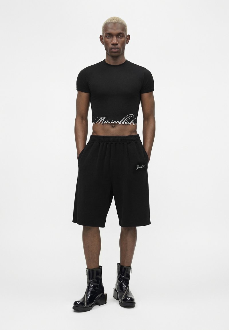 Man with short blonde hair wears a black crop top with "Masallah" text, black shorts, and shiny black ankle boots, standing with hands in pockets.