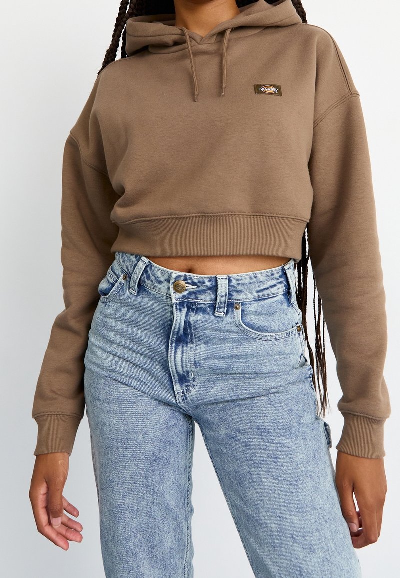 Person som bär en kort brun hoodie med en logo på bröstet och högmidjade ljusblå acid-wash jeans, stående mot en vit bakgrund.