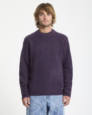Jonge man met verward haar, gekleed in een donkerpaarse gebreide trui en lichtblauwe acid-wash jeans, staande tegen een witte achtergrond.