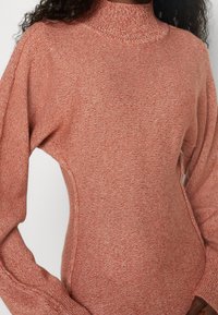 Pull en maille de couleur rouille avec un col haut, des manches longues et des fentes sur les côtés. Texture lisse avec un motif subtil de points.