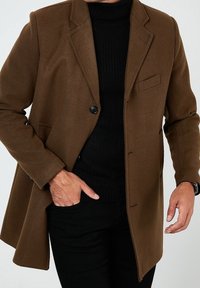 Brauner Wollmischungs-Mantel mit Reverskragen, Einzelknopfverschluss, geradem Schnitt und seitlichen Taschen, getragen über einem schwarzen Rollkragenpullover.