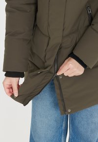 Olivgrön puffjacka med dragkedja, med tryckknappar och elastiska ärmmuddar, visas när den dras upp över blå jeans.