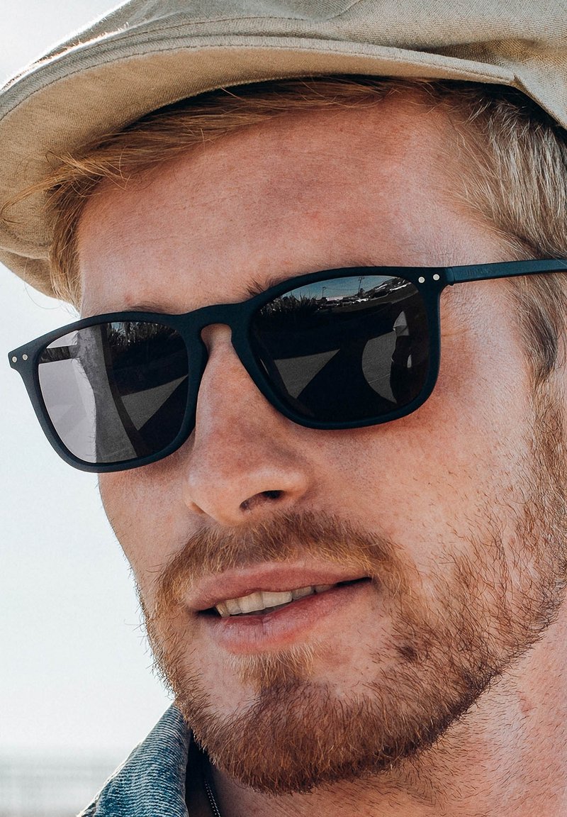 Gafas de sol rectangulares negras con lentes oscuros, acabado mate y pequeños acentos metálicos en la montura, llevadas por una persona con un sombrero de color claro.