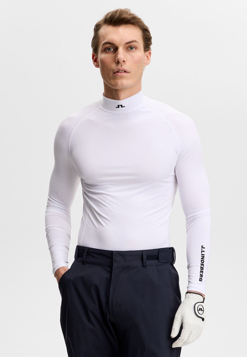Mann trägt ein enganliegendes weißes Langarmshirt mit Logo am Kragen und Ärmel, schwarze Hose und einen weißen Golfhandschuh an seiner rechten Hand.