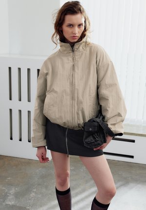 Jeune femme portant une veste beige oversize, une mini-jupe noire avec une fermeture éclair à l'avant, des chaussettes noires hautes et tenant un sac noir à l'intérieur.