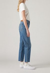 Kvinna som bär en vit t-shirt, högmidjade blå jeans, vita strumpor och vita sneakers, stående mot en enfärgad ljus bakgrund.