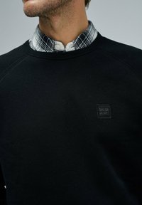 Sudadera negra con un acabado texturizado, con mangas raglán y un pequeño logotipo en relieve. Debajo, se puede ver una camisa de franela a cuadros.