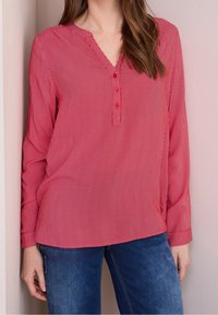 Red and white striped long-sleeve blouse with a V-neck, featuring three buttons at the front and a relaxed fit. Paired with blue jeans.