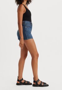 Denim shorts in medium blue met een hoge taille en een rafelrand, gecombineerd met een zwart tanktop en zwarte sandalen met bandjes.