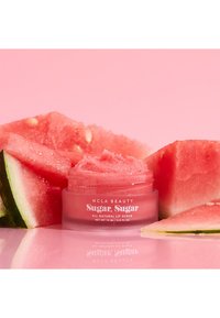 100% naturlig leppeskrubb i et klart glass med rosa lokk, omgitt av vannmelon-skiver på en rosa bakgrunn. Glasset inneholder rosa skrubbtekstur.