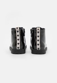 Černé kotníkové boty z lakované kůže s texturovanou podrážkou, na zadní straně s ozdobným bílým páskem s černými srdíčkovými vzory.