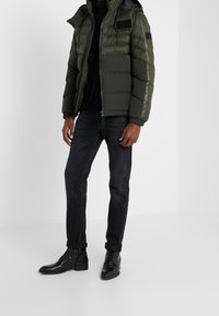 Mand iført en mørk olivengrøn puffjakke, sort turtleneck, mørke optrukne jeans og sorte læder snørebånds støvler.