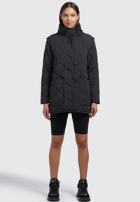 Veste matelassée noire avec un col haut, une fermeture pression et deux poches avant. Portée par-dessus un short noir et des chaussures noires.