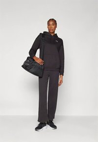 Čierna mikina s kapucňou a zodpovedajúce nohavice vyrobené z mäkkej látky, doplnené textúrovanou čiernou športovou taškou a čiernymi teniskami.