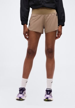 Person trägt beige Sportshorts, weiße Adidas-Socken, lila Laufschuhe und ein schwarzes Langarmshirt, steht vor einem einfarbigen Hintergrund.