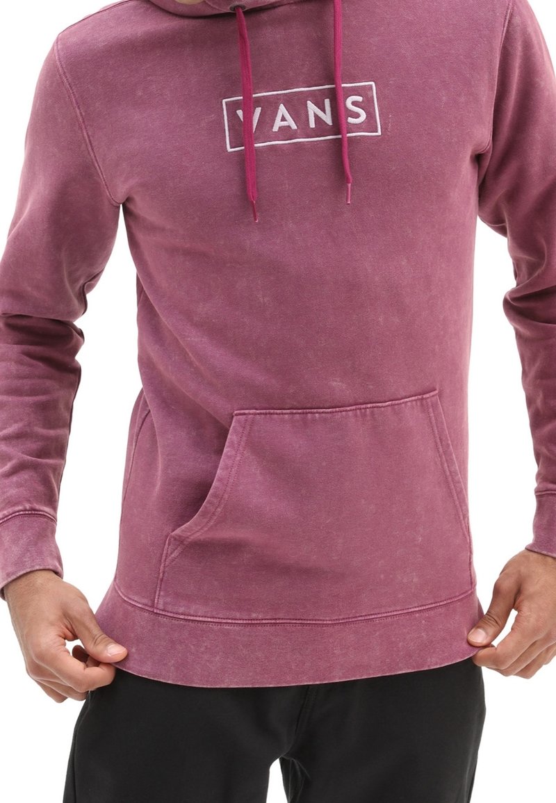 Homme portant un sweat à capuche Vans violet délavé avec une poche avant, ajustant l'ourlet, photographié de la poitrine aux hanches.
