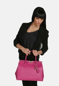 Bolso de cuero rosa con forma estructurada, acentos de hardware dorado y una pequeña etiqueta colgante. Tiene dos asas superiores para llevar.