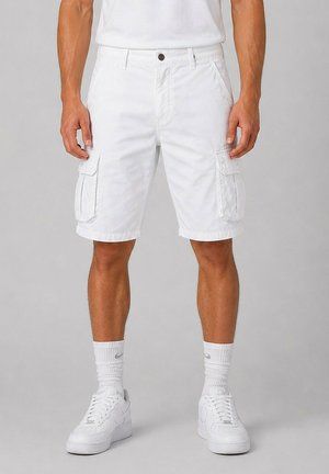 Homme portant un short cargo blanc, des chaussettes blanches avec logo et des baskets blanches, debout devant un fond gris uni.