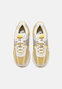 Os ténis Nike Vomero 5 apresentam um upper de malha branca com detalhes amarelos, seções lisas em cinza e uma sola branca almofadada.