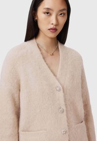 Młoda kobieta z prostymi ciemnymi włosami, ubrana w miękki beżowy sweter z guziczkami ozdobnymi oraz srebrny naszyjnik z złotym zawieszką.