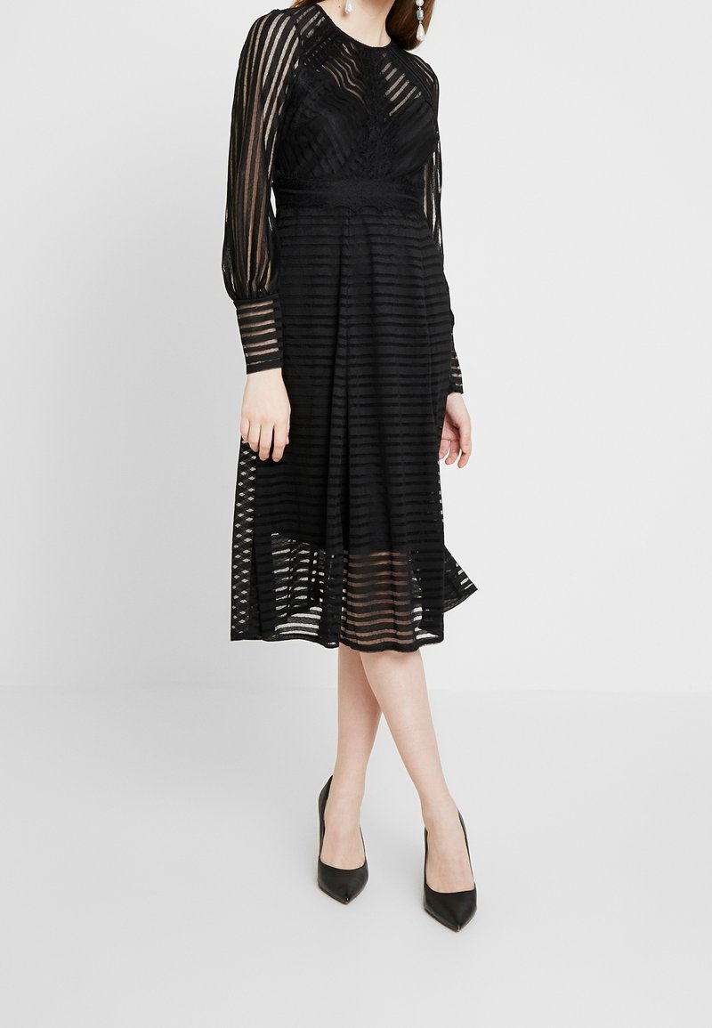 Femme portant une robe midi noire avec des manches longues transparentes rayées et une taille ajustée, associée à des talons aiguilles noirs pointus.