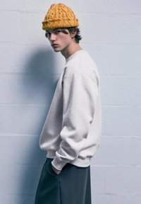 Felpa grigia chiaro con vestibilità rilassata e polsini a costine, abbinata a pantaloni verde scuro. Un berretto di lana lavorato a maglia giallo con texture completa il look.