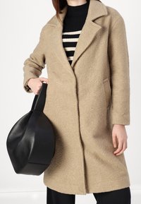 Béžový kabát z vlněné směsi s texturovaným povrchem, klopovými límci s výstřihem a bočními kapsami, doplněný černou koženou taškou s jedinečným, sochařským tvarem.