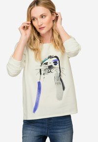 Camicia bianca a maniche lunghe con un disegno colorato di animali illustrati e testo. Abbinata a jeans in denim blu. Tessuto morbido e casual.