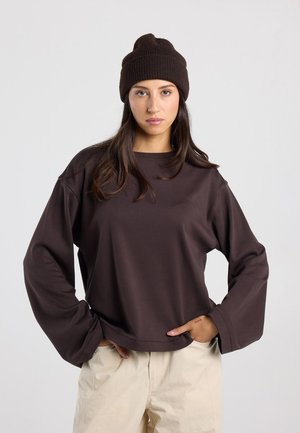 Jeune femme portant un bonnet marron foncé et une chemise à manches longues assortie avec un pantalon beige, debout les mains dans les poches.