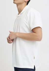 Hvit polotrøye med klassisk krage, korte ermer og sideslisser. Stoffet fremstår mykt med en teksturert overflate og subtil logo.