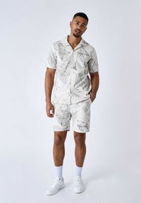 Camisa ligera de manga corta con botones y pantalones cortos a juego. Ambos presentan una base blanca con patrones de hojas tropicales en gris. Se lleva con zapatillas blancas.